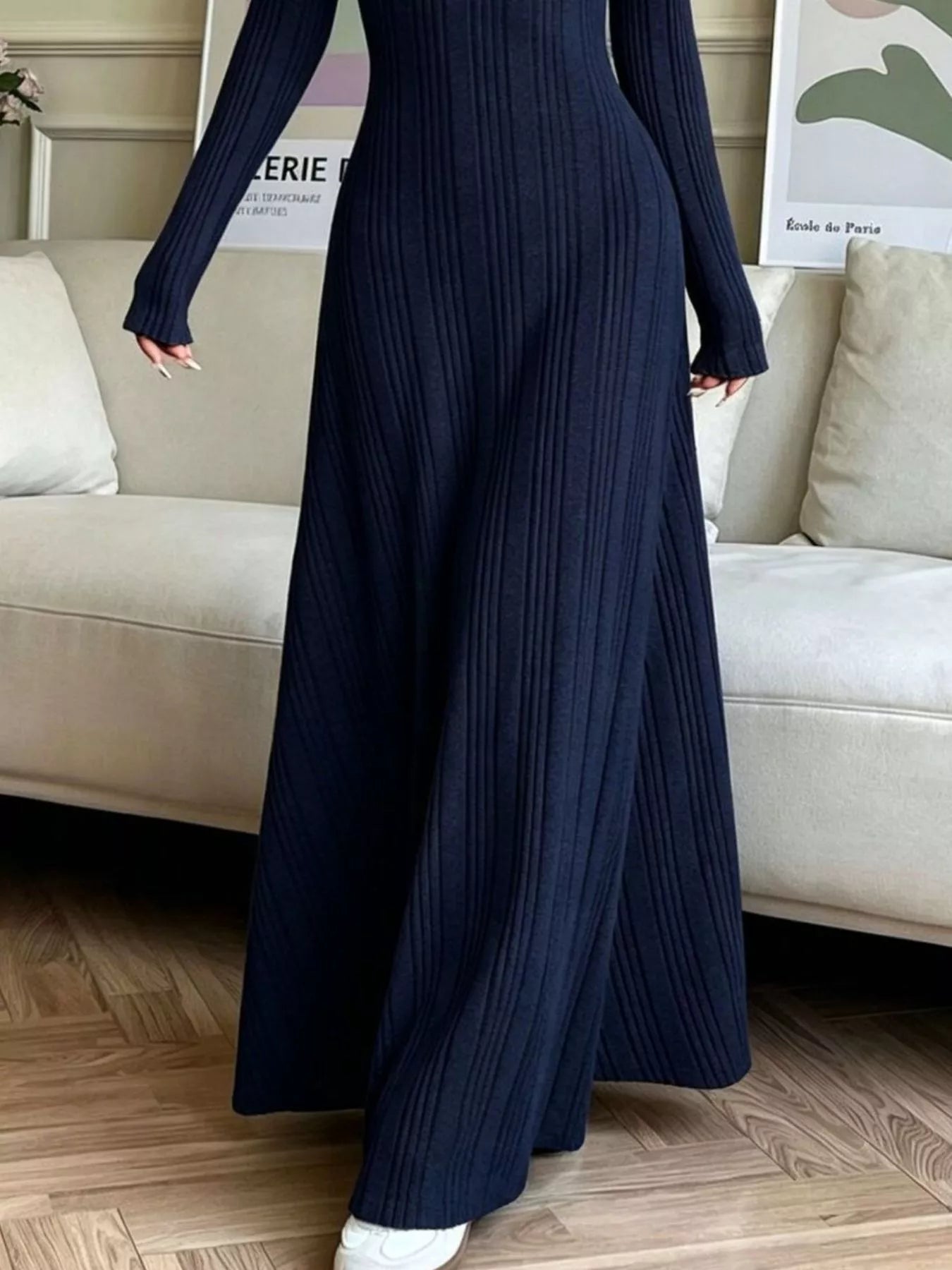 NEWYORKLISTED STYLE COLLECTION: A person wearing a long-sleeve, floor-length navy blue ribbed knit maxi dress, standing in a living room with a beige sofa and wood floor.