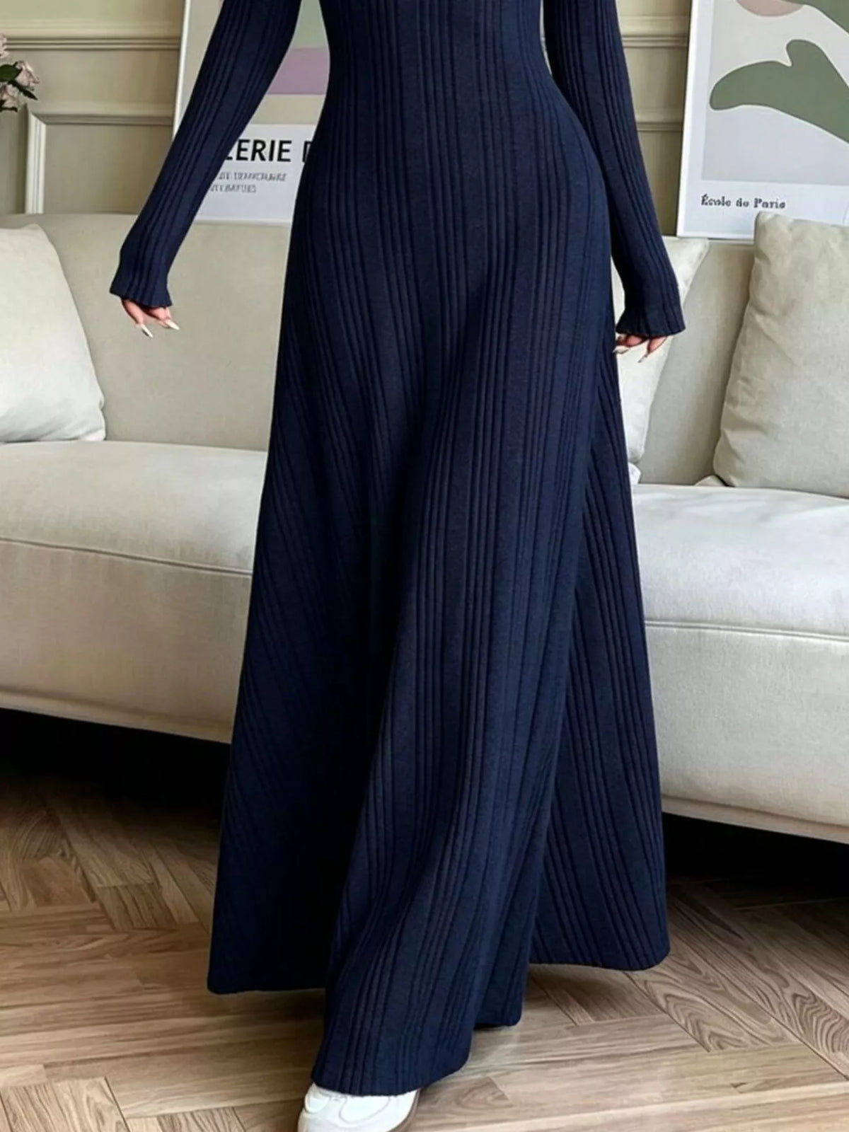 NEWYORKLISTED STYLE COLLECTION: A person wearing a long-sleeve, floor-length navy blue ribbed knit maxi dress, standing in a living room with a beige sofa and wood floor.