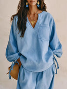 NEWYORKLISTED STYLE COLLECTION: A woman wearing a light blue oversized long-sleeve top with a V-neck, chest pocket, and side tie details, paired with matching light blue bottoms. She is accessorized with gold jewelry and blue earrings against a neutral beige background.