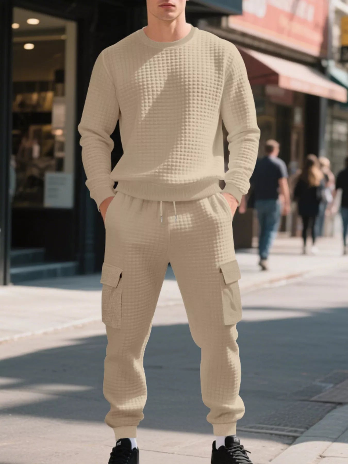 NEWYORKLISTED ATLAS MEN'S COLLECTION: A man wearing a matching beige waffle-knit sweatshirt and cargo jogger pants stands on a city sidewalk, with blurred storefronts and pedestrians in the background.