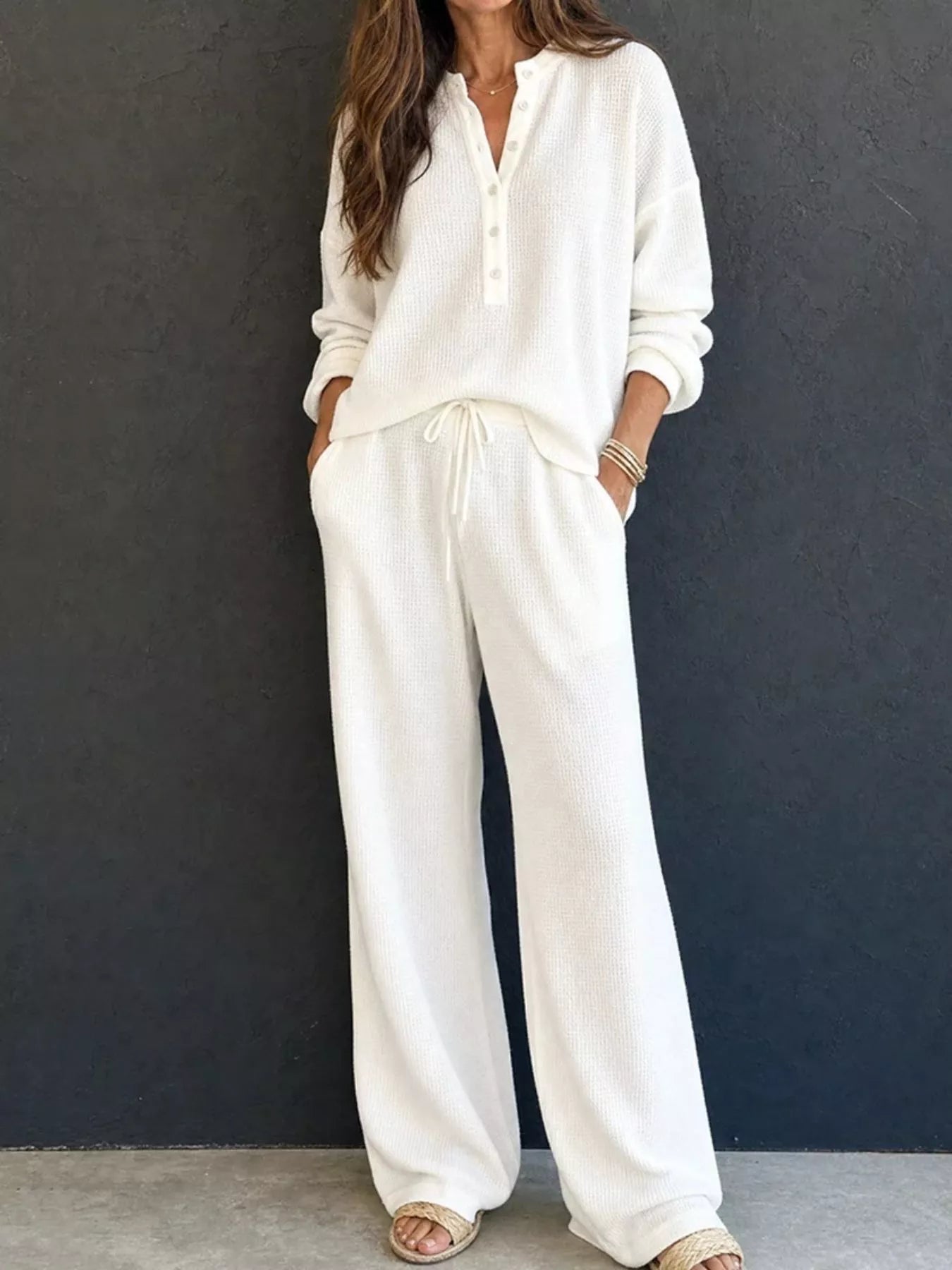 NEWYORKLISTED STYLE & COMFORT COLLECTION: A woman wearing a matching off-white knit lounge set consisting of a long-sleeve henley top and wide-leg drawstring pants, standing against a dark textured wall.
