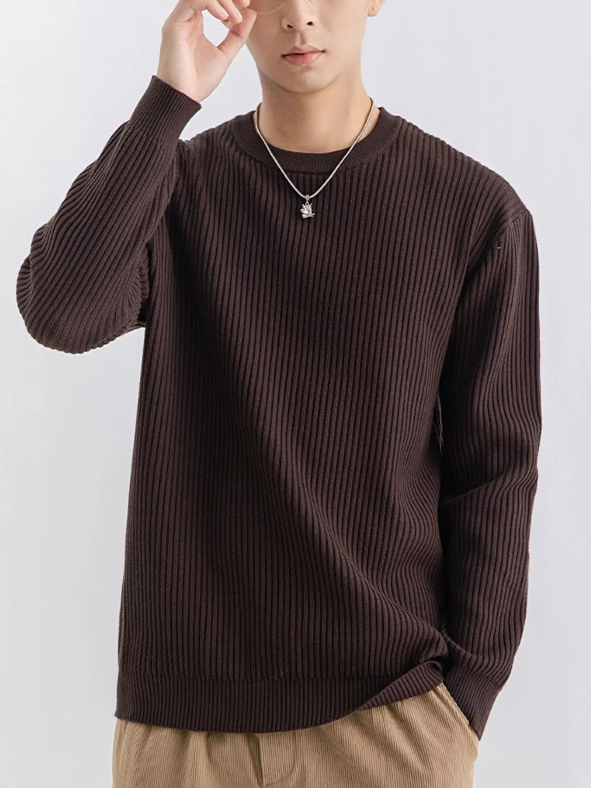 NEWYORKLISTED ATLAS MEN'S COLLECTION: A man wearing a dark brown ribbed knit crewneck sweater, paired with beige pants and a silver necklace, against a plain light gray background.