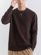 NEWYORKLISTED ATLAS MEN'S COLLECTION: A man wearing a dark brown ribbed knit crewneck sweater, paired with beige pants and a silver necklace, against a plain light gray background.