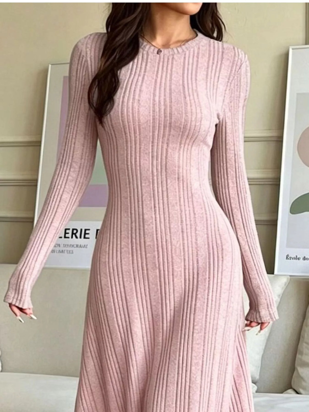 NEWYORKLISTED STYLE COLLECTION: A woman wearing a long-sleeve, ribbed knit midi dress in a soft dusty pink color, standing in an indoor setting with framed wall art in the background.