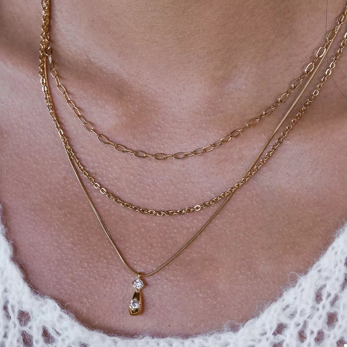 NYLD Glamour Collection by NEWYORKLISTED: Close-up of a person wearing two gold necklaces with a delicate pendant on a soft background.