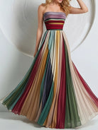 NEWYORKLISTED STYLE COLLECTION: A woman wearing a strapless A-line maxi dress with a multicolor striped bodice and flowing pleated skirt in shades of green, red, beige, and blue, posed against a white background with a circular textured wall.