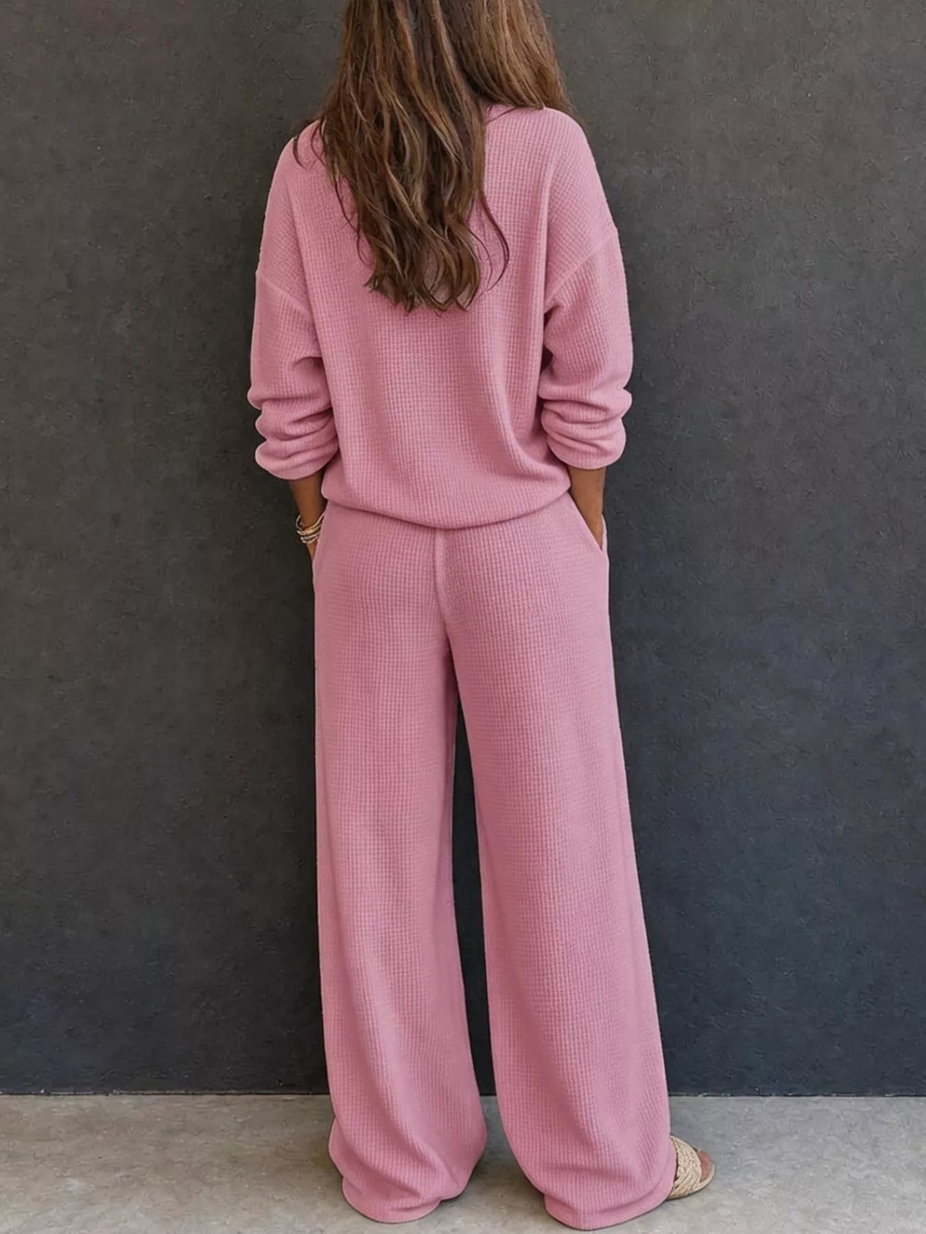 NEWYORKLISTED STYLE & COMFORT COLLECTION: A woman viewed from behind wearing a matching dusty pink knit lounge set consisting of a long-sleeve top and wide-leg pants, standing against a dark gray textured wall.