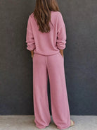 NEWYORKLISTED STYLE & COMFORT COLLECTION: A woman viewed from behind wearing a matching dusty pink knit lounge set consisting of a long-sleeve top and wide-leg pants, standing against a dark gray textured wall.