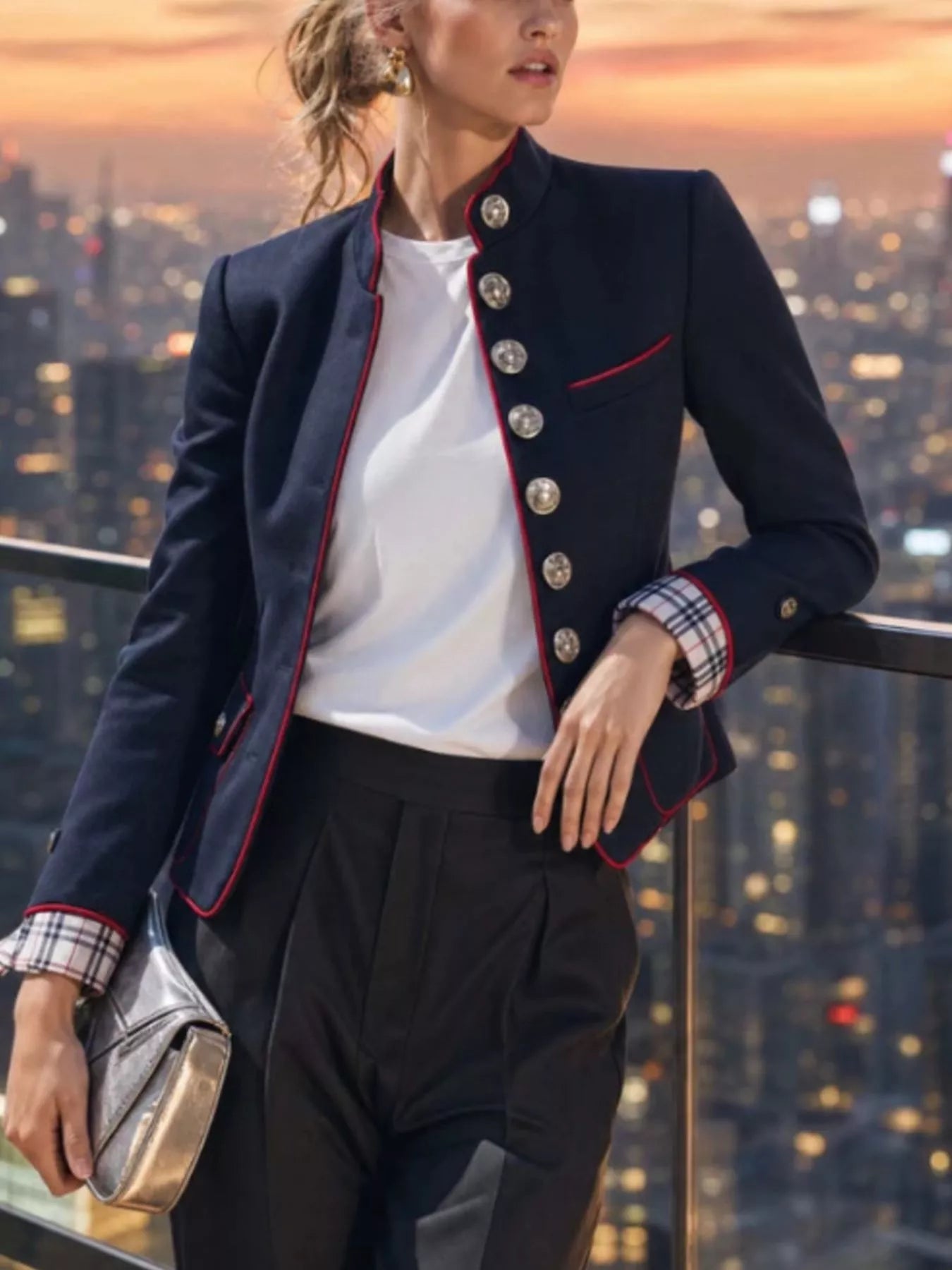 NEWYORKLISTED STYLE COLLECTION:  A woman models a navy blue blazer with red piping and silver buttons, worn over a white top and black trousers, set against a city skyline at sunset.