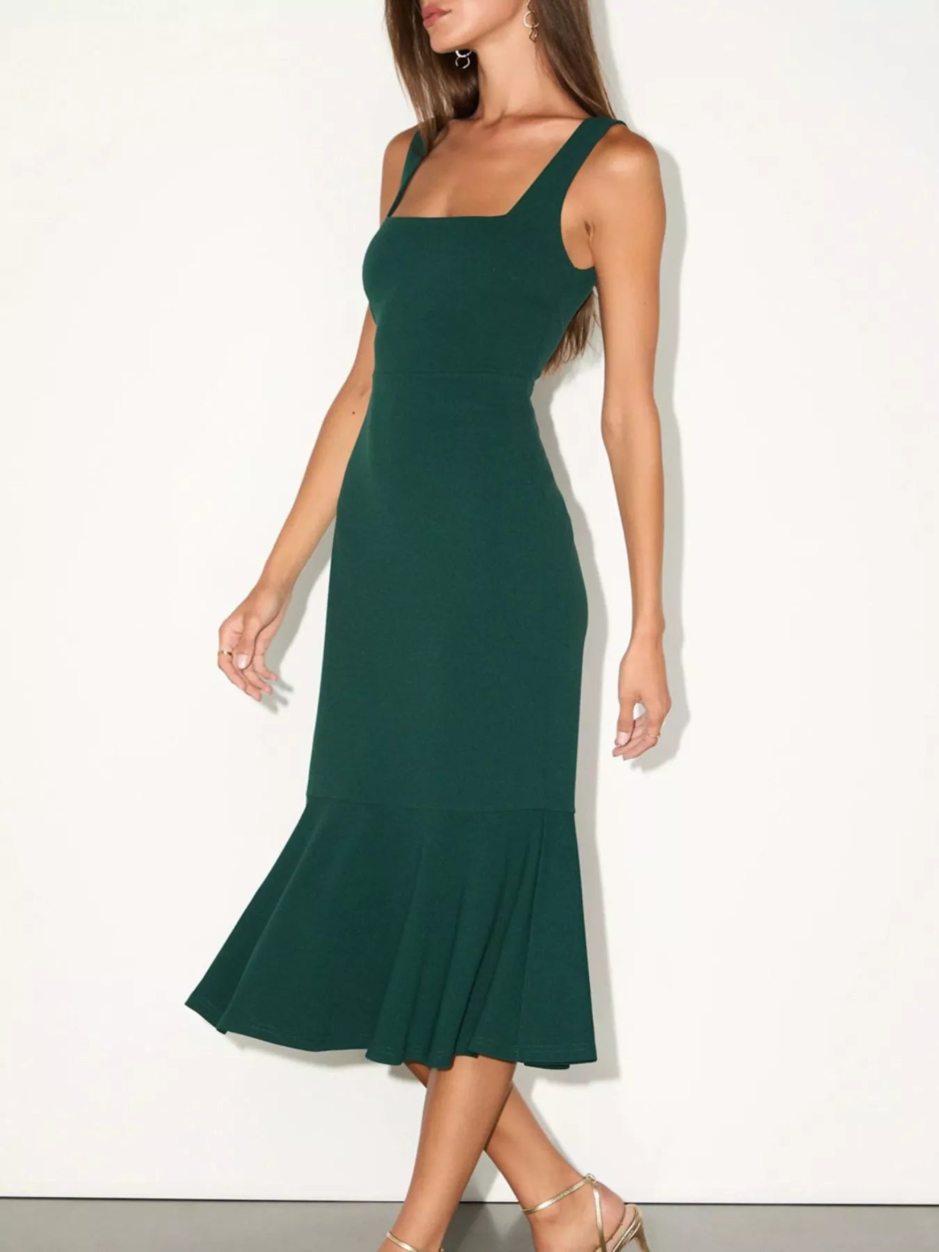 NEWYORKLISTED STYLE COLLECTION: A woman modeling a dark green midi dress with a square neckline, wide shoulder straps, and a flared ruffled hem, photographed against a plain off-white studio background.