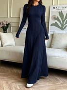 NEWYORKLISTED STYLE COLLECTION: A woman wearing a navy blue long-sleeve maxi dress with a vertical pleated texture, standing in a living room setting with a beige sofa and wooden floor.