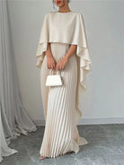 NEWYORKLISTED STYLE COLLECTION: Woman wearing a beige cape over a pleated dress holding a white clutch against a plain background