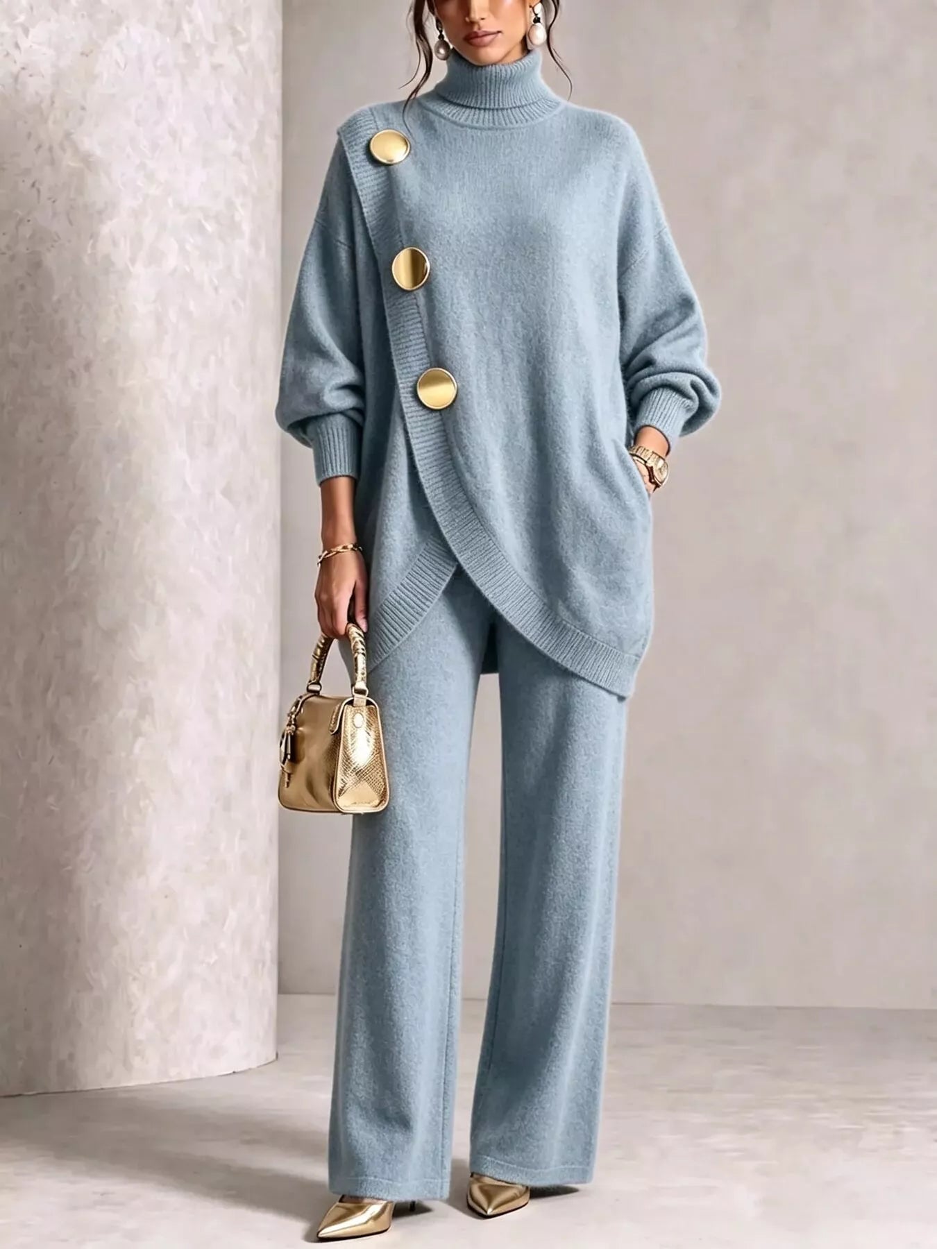NEWYORKLISTED STYLE COLLECTION:  A woman models a light blue knit two-piece set consisting of an asymmetrical turtleneck sweater with large gold buttons and matching wide-leg trousers. She is holding a small metallic gold handbag and wearing gold pointed shoes, set against a neutral textured beige background.