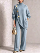 NEWYORKLISTED STYLE COLLECTION:  A woman models a light blue knit two-piece set consisting of an asymmetrical turtleneck sweater with large gold buttons and matching wide-leg trousers. She is holding a small metallic gold handbag and wearing gold pointed shoes, set against a neutral textured beige background.