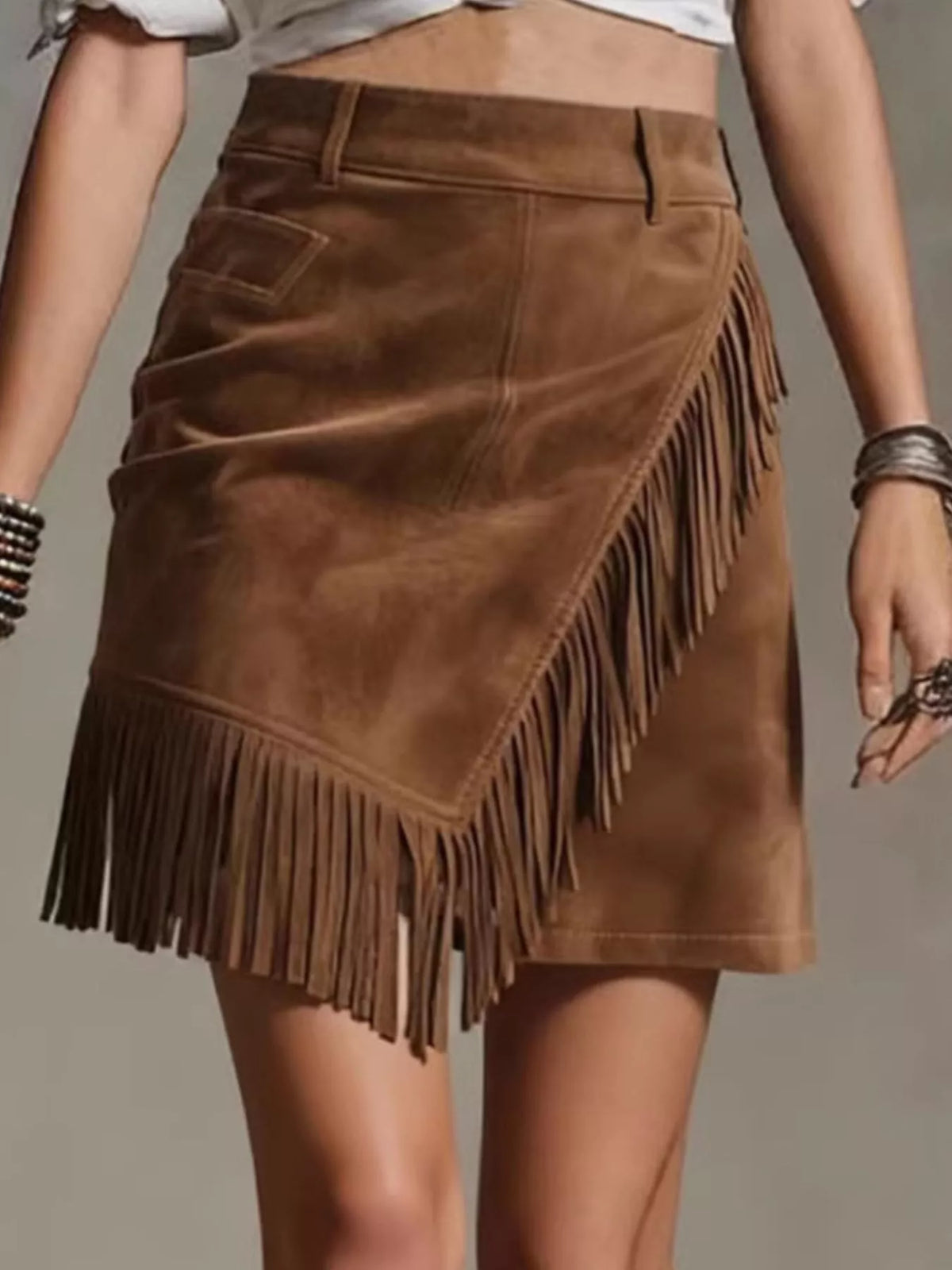 NEWYORKLISTED STYLE COLLECTION: A brown suede mini skirt with asymmetrical fringe detailing, shown on a model from the waist down against a neutral gray background.
