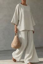 NEWYORKLISTED STYLE COLLECTION: A model wearing a light beige linen two-piece set consisting of a loose-fitting short-sleeve top and matching wide-leg pants, holding a woven straw handbag against a textured gray wall.
