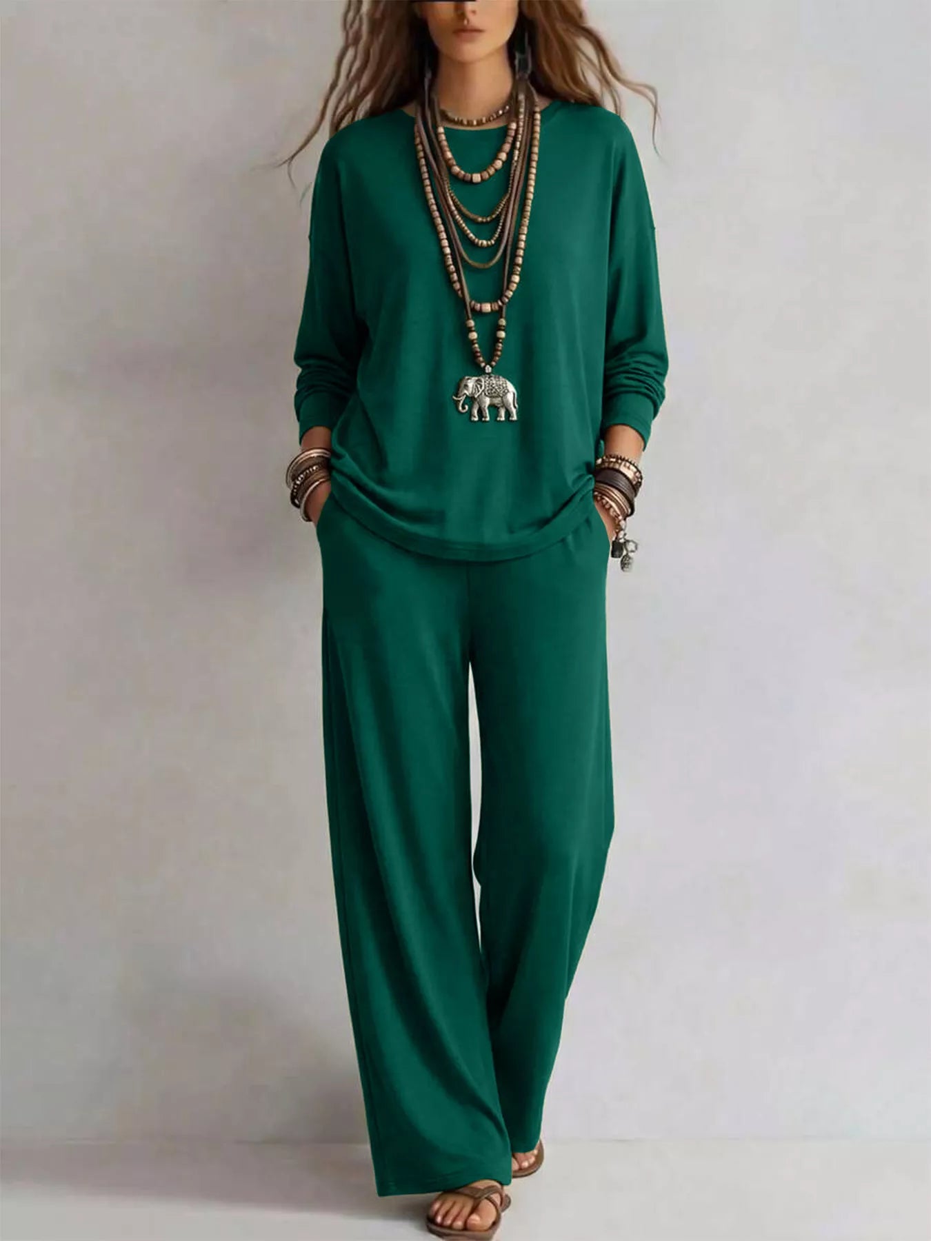 NEWYORKLISTED STYLE COLLECTION: A woman modeling a matching dark green long-sleeve top and wide-leg pants set, accessorized with layered necklaces, bracelets, and sandals, against a plain light gray background.