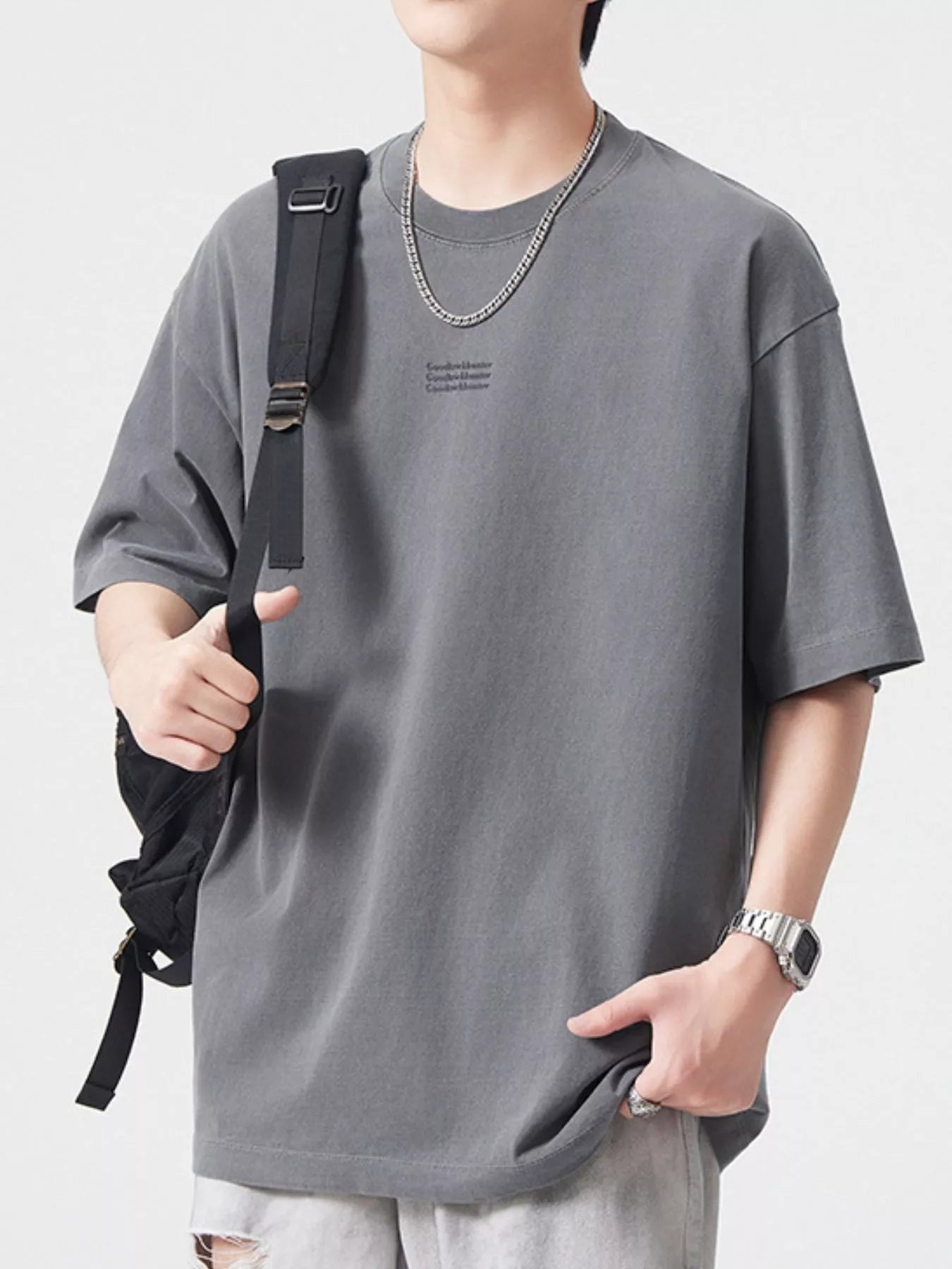 NEWYORKLISTED ATLAS MEN's COLLECTION: Person wearing a gray t-shirt with a small logo, holding a black backpack on a light gray background