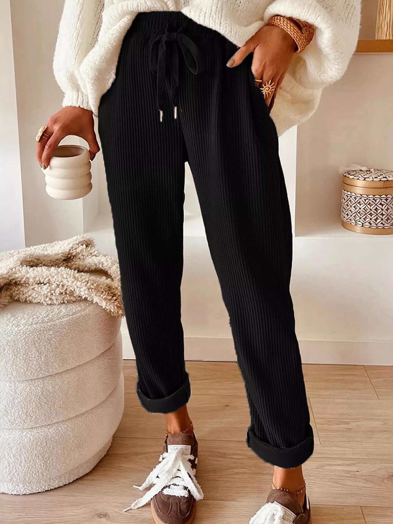 NEWYORKLISTED STYLE COLLECTION: Person wearing black ribbed pants and a white sweater, holding a ceramic cup indoors.