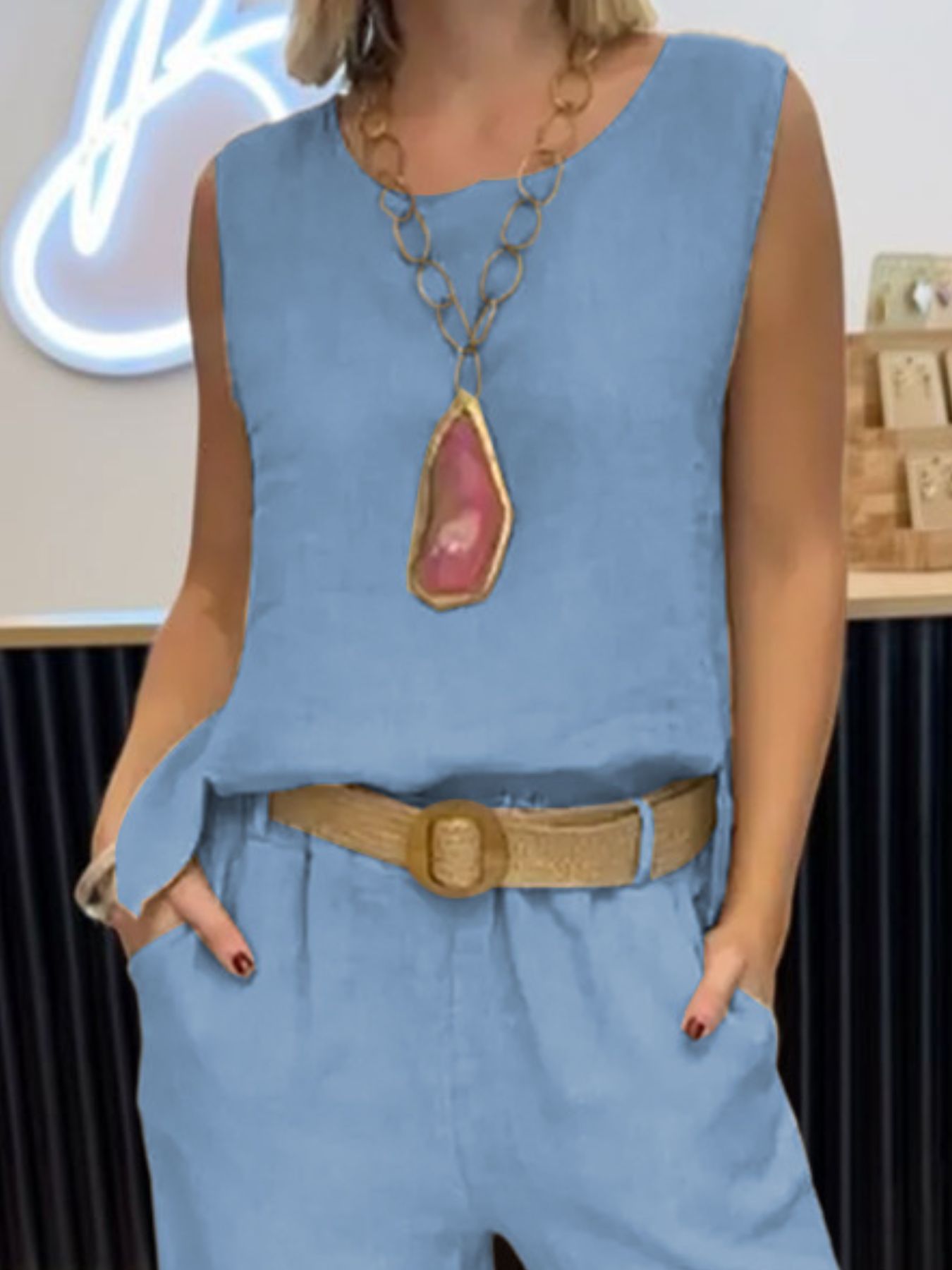 NEWYORKLISTED COMFORT COLLECTION: Person wearing a blue sleeveless top and pants with a large necklace, standing indoors.
