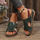 NEWYORKLISTED SHOE COLLECTION: A pair of dark green leather slide sandals with cutout details, white contrast stitching, and a brown heel strap, shown on a model's feet on a rustic wooden floor.