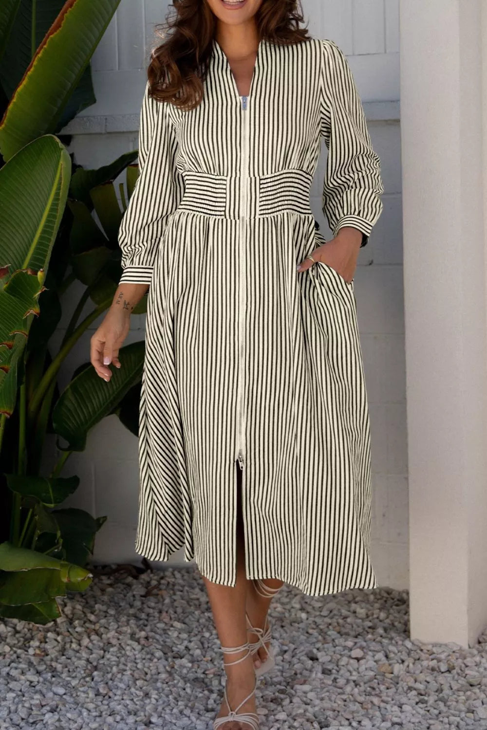 NEWYORKLISTED STYLE COLLECTION: Woman wearing a striped dress standing outdoors with plants and a white wall in the background