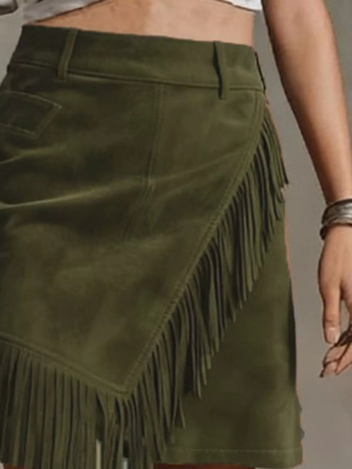 NEWYORKLISTED STYLE COLLECTION: An olive green suede skirt with long fringe trim, shown on a model. The skirt features an asymmetrical design with a side pocket and a belt loop.