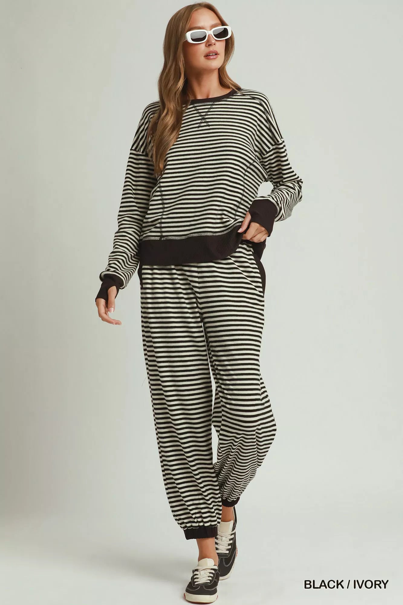NEWYORKLISTED COMFORT COLLECTION: A woman modeling a matching black and ivory striped lounge set consisting of a long-sleeve top and jogger pants, styled with white sunglasses and black sneakers against a light gray studio background.