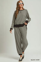 NEWYORKLISTED COMFORT COLLECTION: A woman modeling a matching black and ivory striped lounge set consisting of a long-sleeve top and jogger pants, styled with white sunglasses and black sneakers against a light gray studio background.