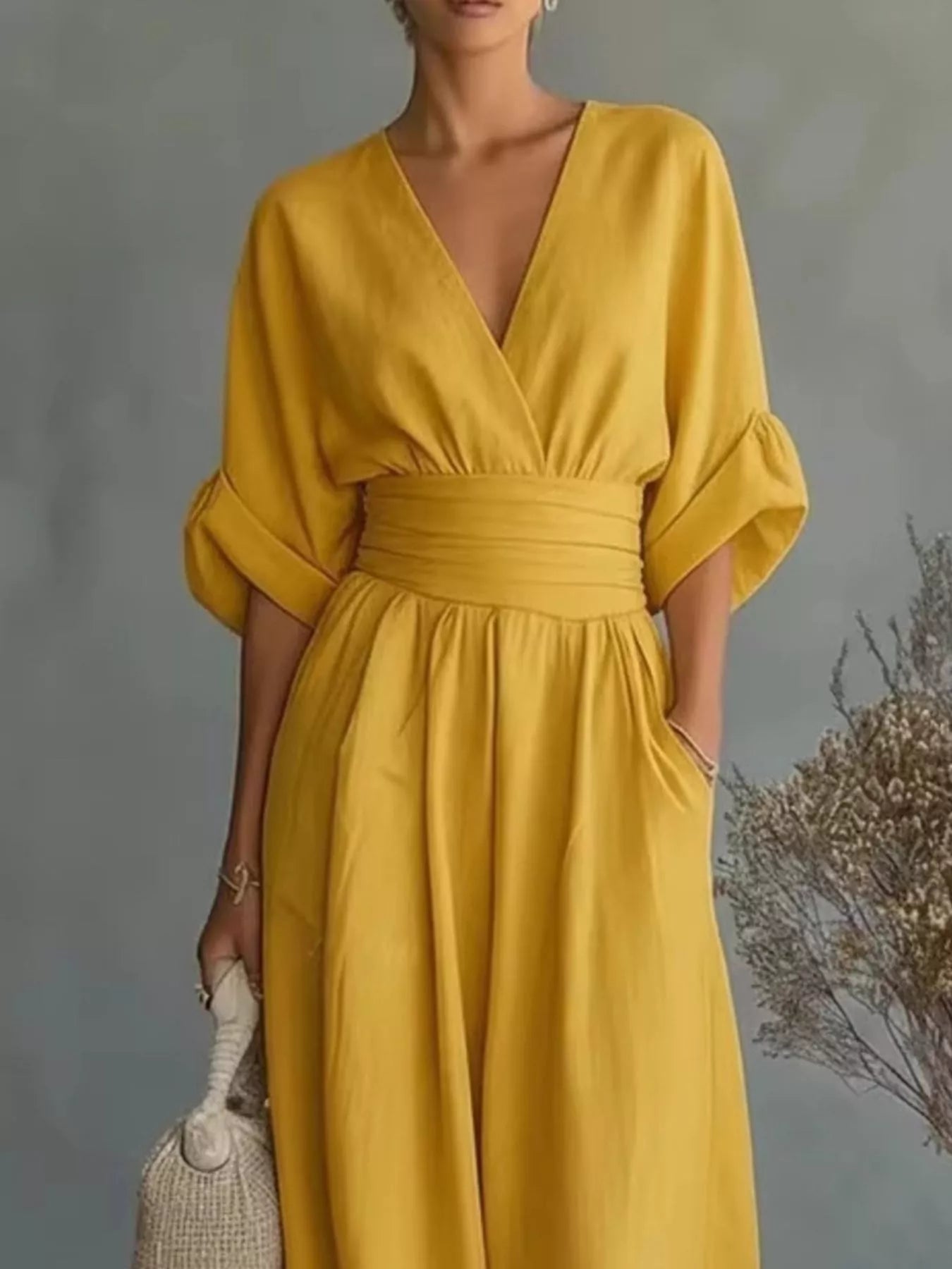 NEWYORKLISTED STYLE COLLECTION: A woman wearing a mustard yellow midi dress with a V-neckline, wide elbow-length sleeves, a matching fabric waist belt, and side pockets, holding a woven cream handbag against a gray studio backdrop with dried floral decor.