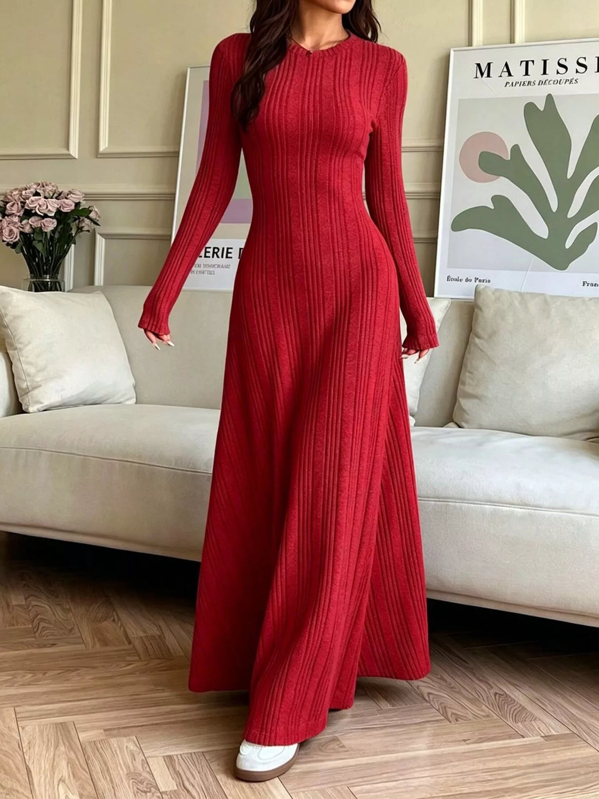 NEWYORKLISTED STYLE COLLECTION: A woman wearing a long-sleeved, floor-length red ribbed knit maxi dress, standing in a living room setting with a beige sofa and framed art in the background.