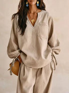 NEWYORKLISTED STYLE COLLECTION: A woman wearing a beige linen-like two-piece set with a long-sleeve V-neck top featuring a chest pocket and matching wide-leg pants, styled with gold jewelry and a straw bag against a neutral beige background.