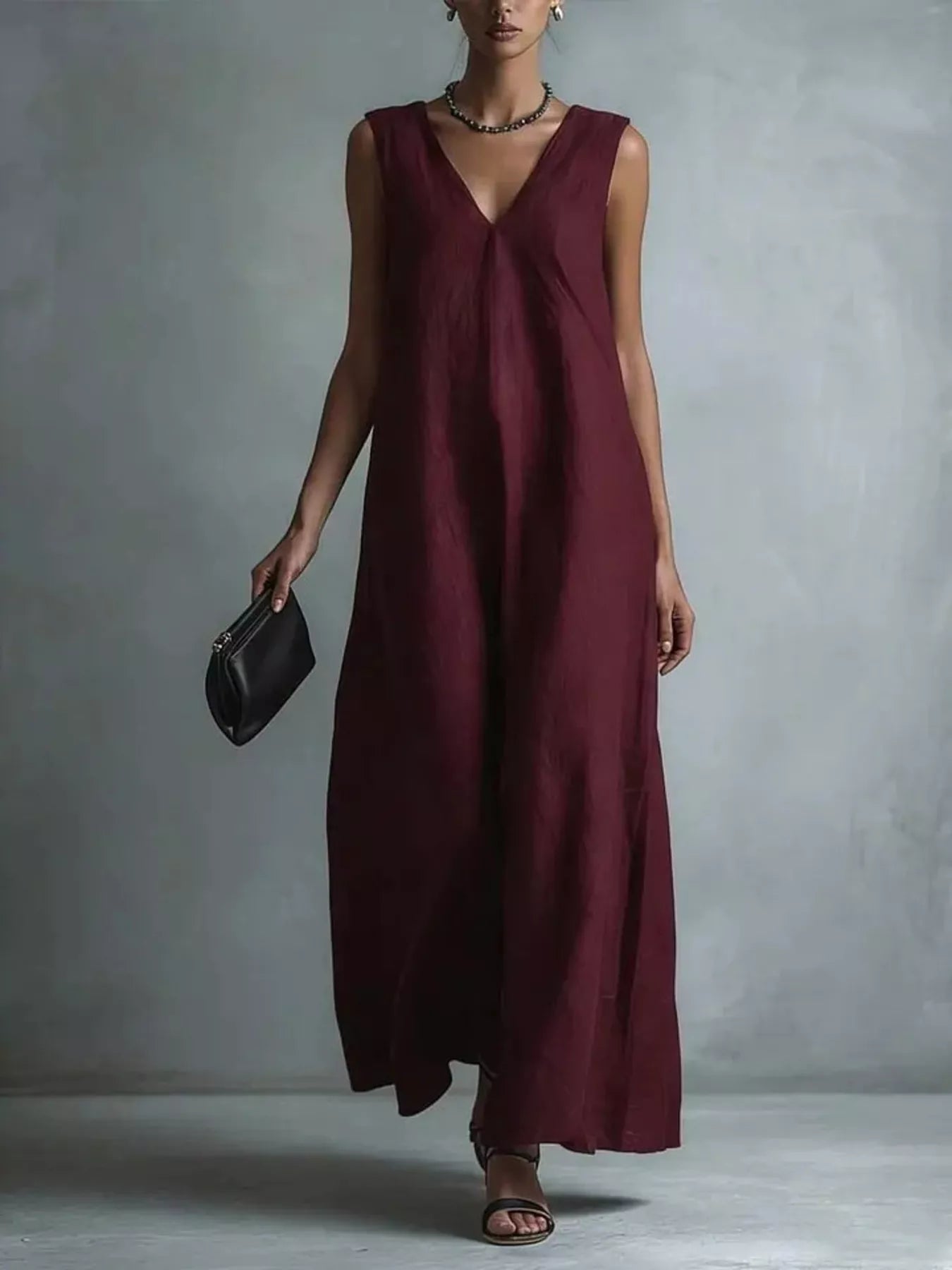NEWYORKLISTED STYLE COLLECTION: A woman models a sleeveless, deep burgundy maxi dress with a V-neckline, holding a small black clutch against a textured gray studio backdrop.