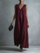 NEWYORKLISTED STYLE COLLECTION: A woman models a sleeveless, deep burgundy maxi dress with a V-neckline, holding a small black clutch against a textured gray studio backdrop.
