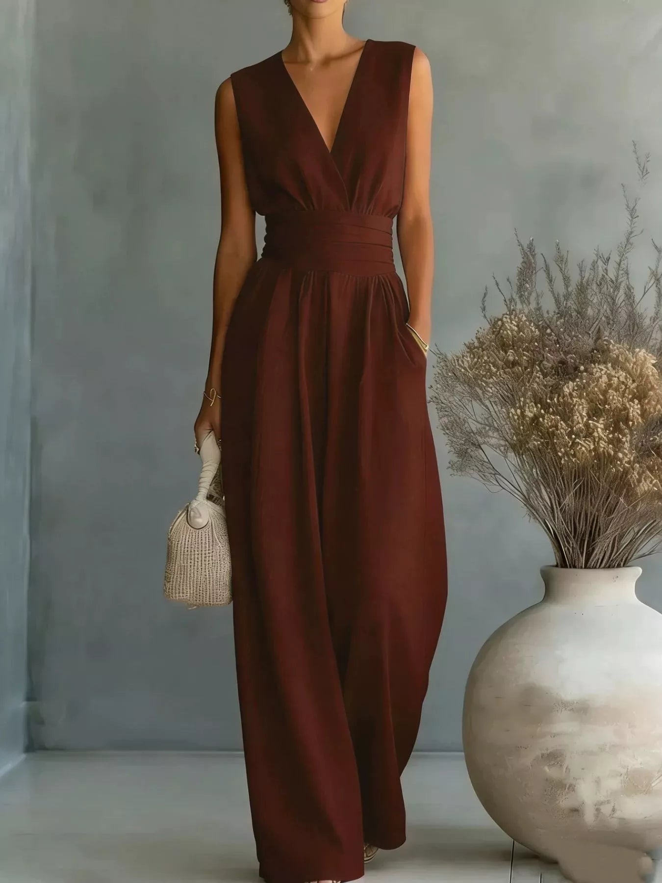 NEWYORKLISTED STYLE COLLECTION: Woman wearing a brown jumpsuit holding a small bag in a minimalistic setting with a vase of dried flowers.