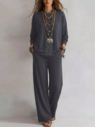 NEWYORKLISTED STYLE COLLECTION: A woman modeling a dark gray two-piece lounge set consisting of a long-sleeve top and matching wide-leg pants, styled with layered gold necklaces and bracelets against a neutral studio background.