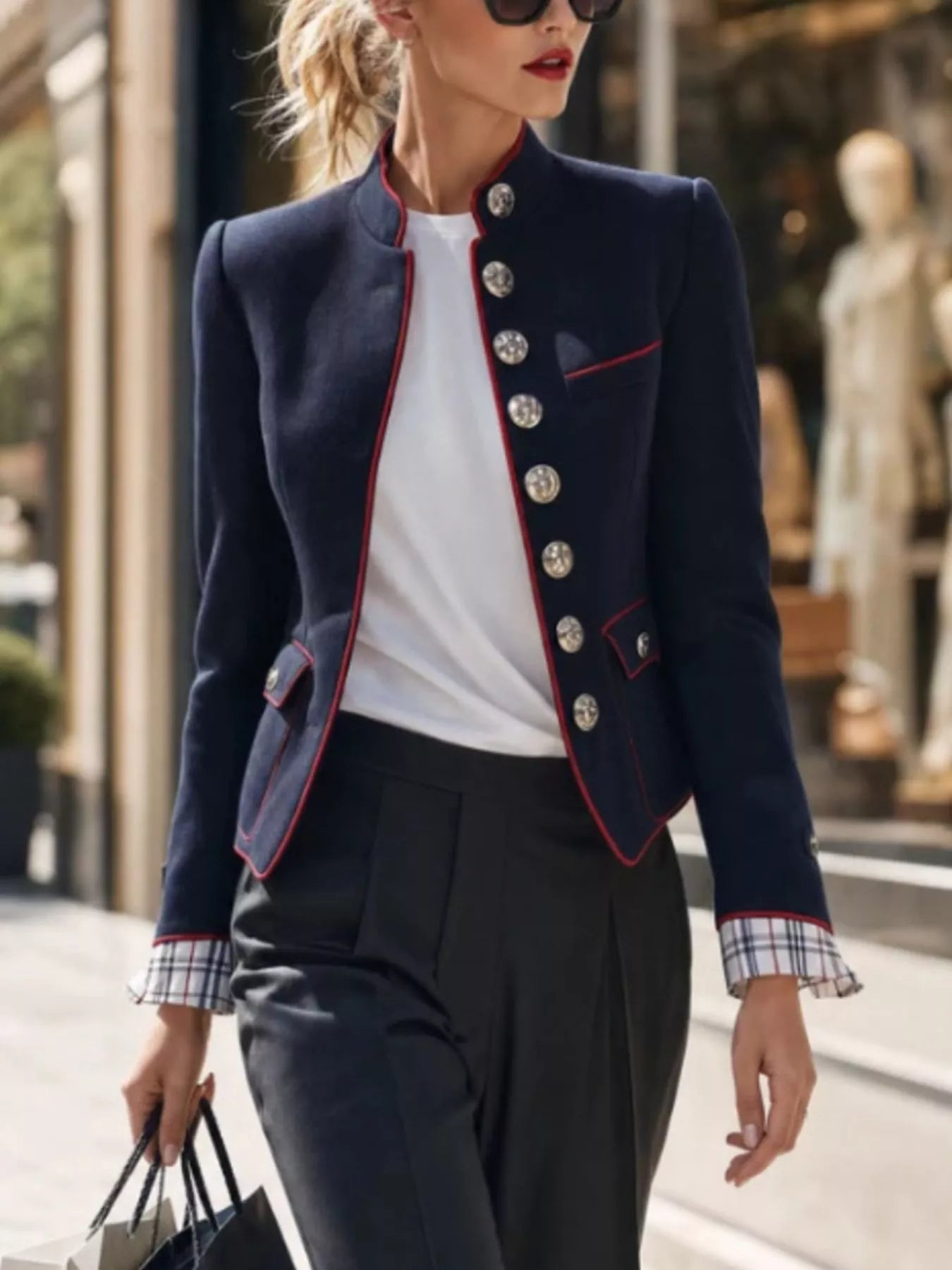 NEWYORKLISTED STYLE COLLECTION: A woman walking outdoors wearing a navy blazer with red piping and silver buttons, a white top, dark trousers, and sunglasses, holding a shopping bag.