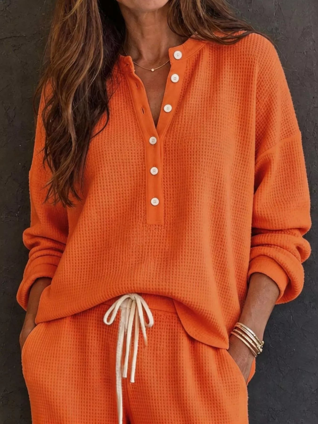 NEWYORKLISTED STYLE & COMFORT COLLECTION: A woman wearing a bright orange waffle-knit two-piece loungewear set, featuring a long-sleeve henley top with white buttons and matching drawstring pants, set against a dark textured wall.