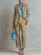 NEWYORKLISTED STYLE COLLECTION: A model wears a matching plaid blazer and trousers set in green, orange, and beige tones, styled with a bright blue shirt, a blue handbag, and blue strappy heels against a textured gray studio backdrop.