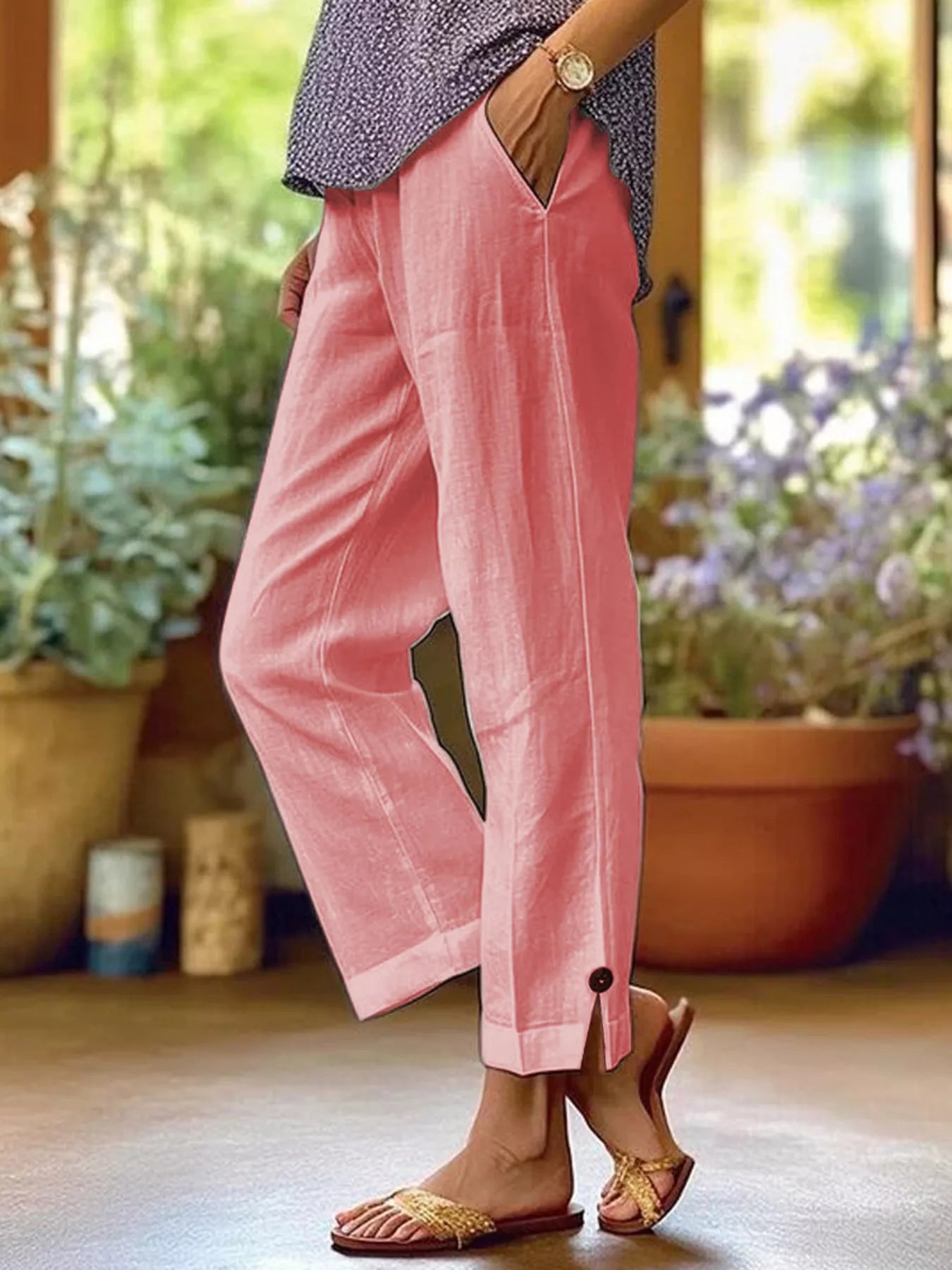 NEWYORKLISTED STYLE COLLECTION: A person wearing loose pink cropped pants with a button detail at the hem, paired with a patterned top and sandals, standing in a sunlit indoor setting with potted plants in the background.