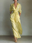 NEWYORKLISTED STYLE COLLECTION: A woman models a flowing pale yellow wrap-style maxi dress with a deep V-neck, short sleeves, side pockets, and a self-tie waist, photographed against a neutral textured wall.