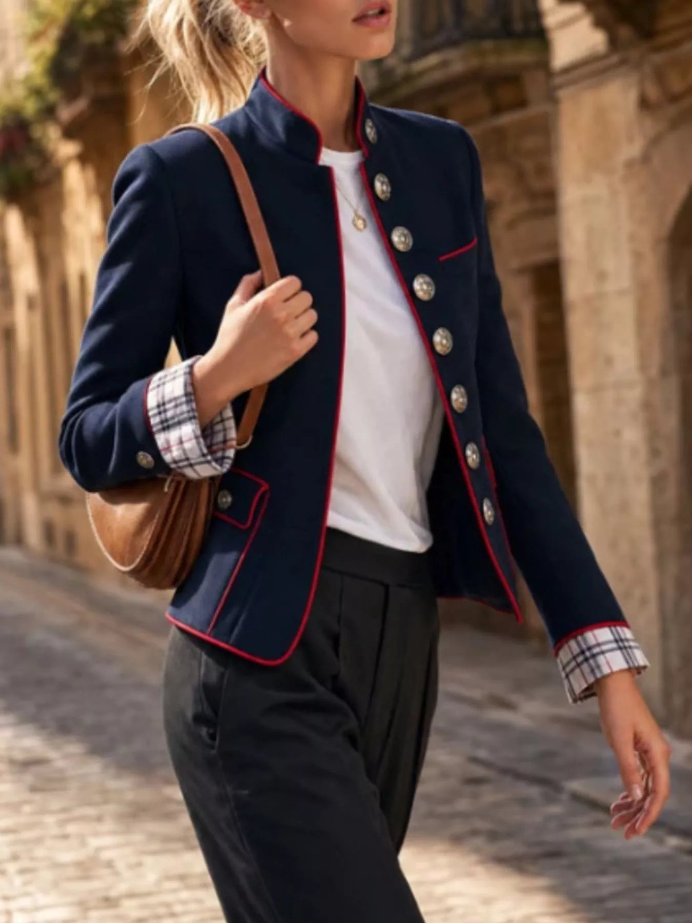 NEWYORKLISTED STYLE COLLECTION:  A woman wearing a navy blue military-style blazer with red piping, silver buttons, and a plaid cuff, paired with a white top and dark trousers, carrying a brown shoulder bag in an outdoor stone alleyway.