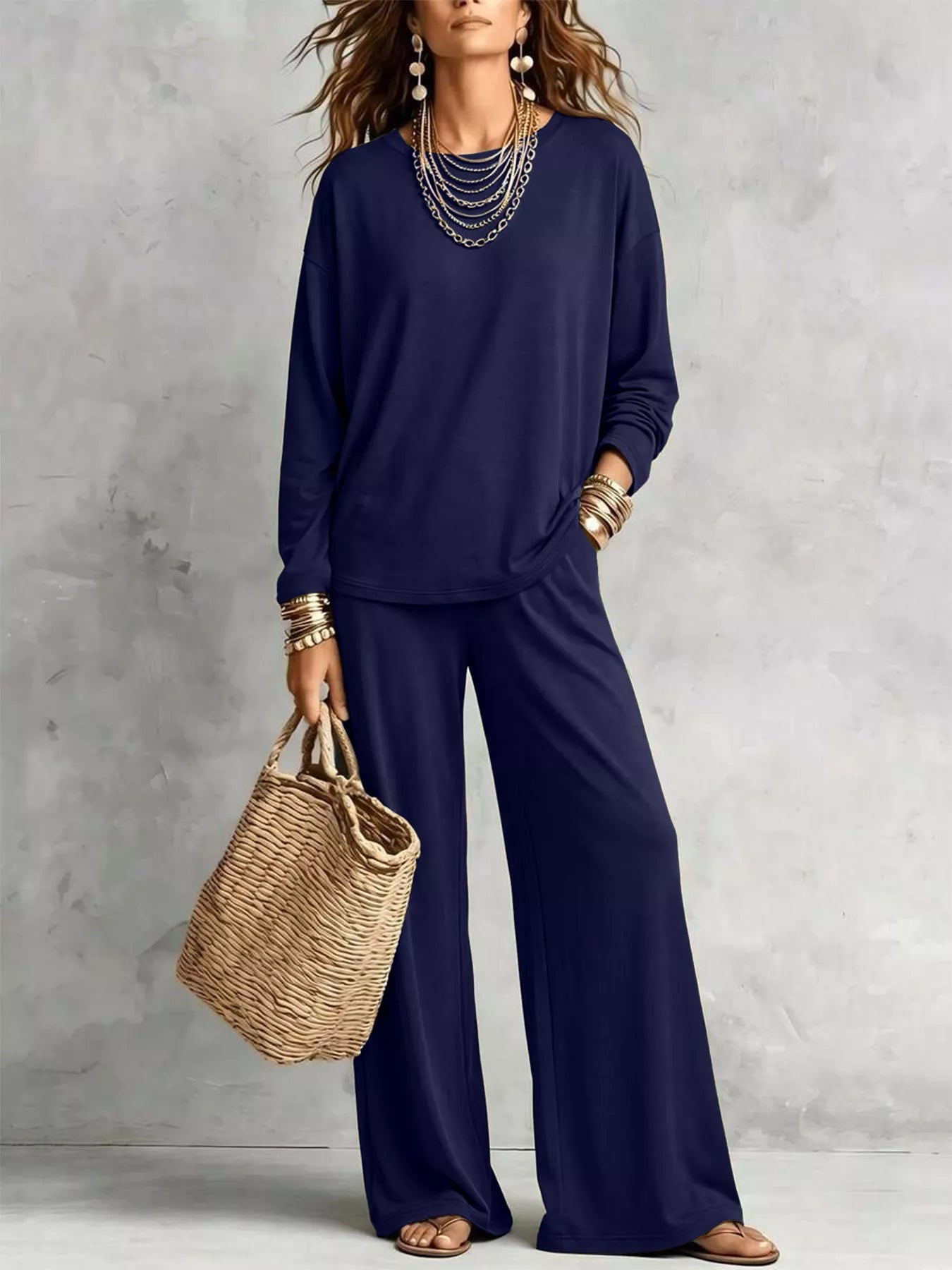 NEWYORKLISTED STYLE COLLECTION: A woman models a matching navy blue lounge set consisting of a long-sleeve top and wide-leg pants, styled with layered silver necklaces, gold bracelets, and a woven straw tote bag against a textured gray wall.