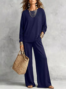NEWYORKLISTED STYLE COLLECTION: A woman models a matching navy blue lounge set consisting of a long-sleeve top and wide-leg pants, styled with layered silver necklaces, gold bracelets, and a woven straw tote bag against a textured gray wall.