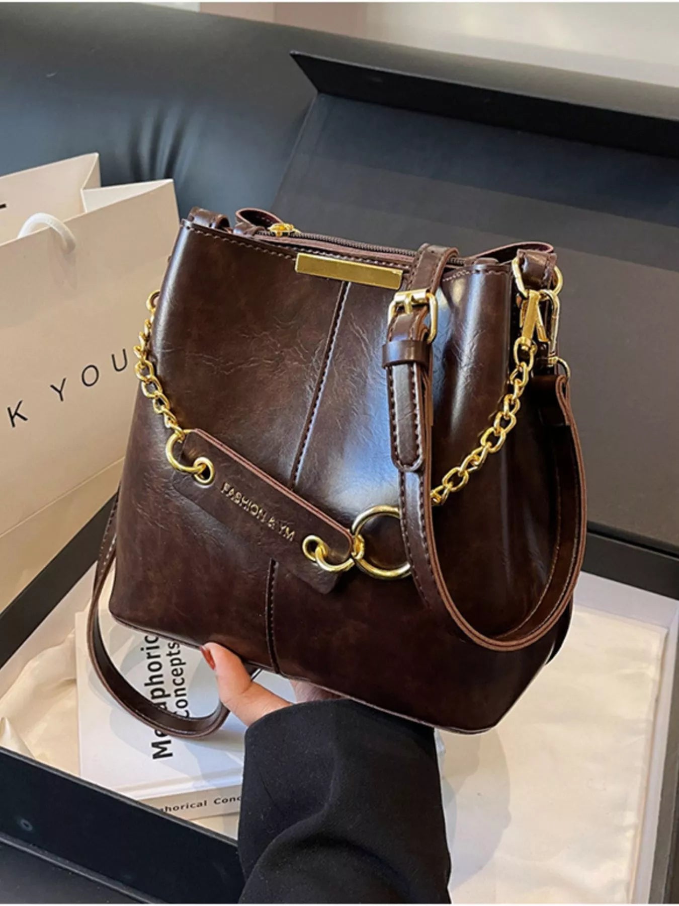 Brown leather handbag with gold chain details held by a person, on a white surface. New York Listed Style Collection