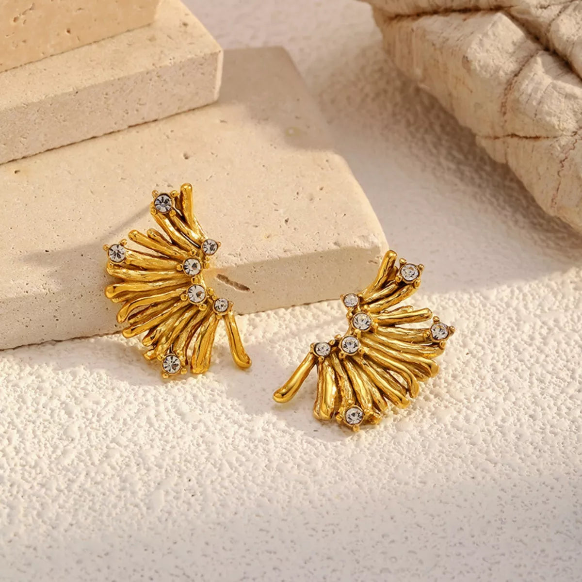 NYLD Glamour Collection by NEWYORKLISTED: Gold earrings with gemstones on a textured beige surface
