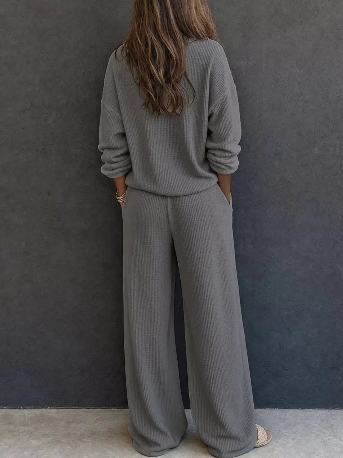 NEWYORKLISTED STYLE & COMFORT COLLECTION: A woman viewed from behind wearing a matching gray ribbed knit lounge set consisting of a long-sleeve top and wide-leg pants, standing against a dark gray textured wall.