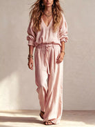 NEWYORKLISTED STYLE COLLECTION: A woman modeling a light pink two-piece linen set consisting of a long-sleeve V-neck top and matching wide-leg trousers, styled with layered necklaces and bracelets against a textured beige wall.
