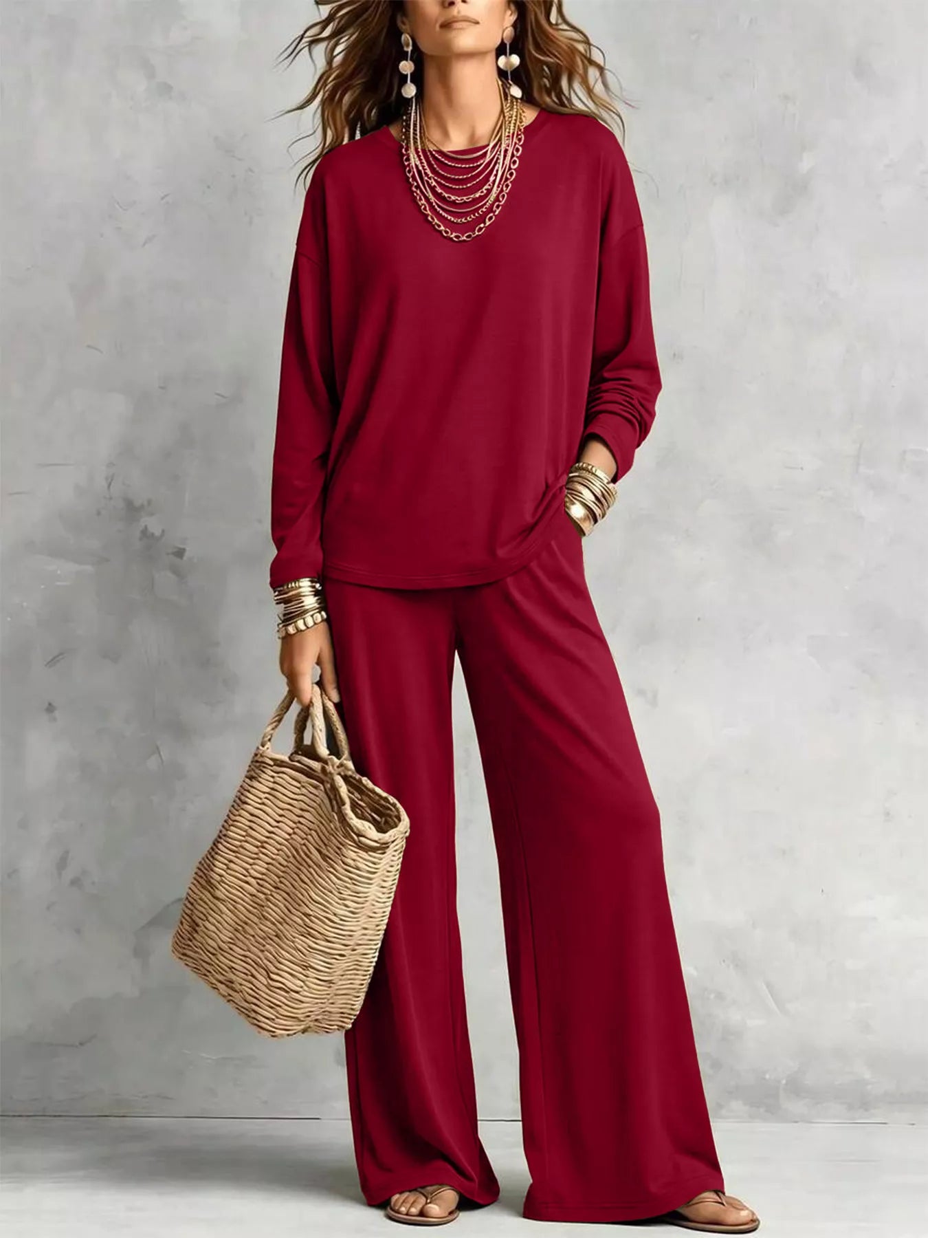 NEWYORKLISTED STYLE COLLECTION: A woman models a matching burgundy long-sleeve top and wide-leg pants set, styled with layered gold necklaces, bracelets, and a straw tote bag against a textured gray wall.