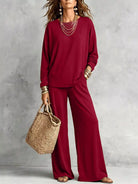 NEWYORKLISTED STYLE COLLECTION: A woman models a matching burgundy long-sleeve top and wide-leg pants set, styled with layered gold necklaces, bracelets, and a straw tote bag against a textured gray wall.